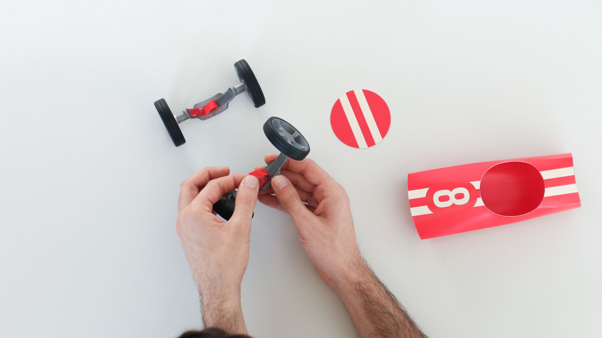 Hands snapping wheels onto a Bendit car beside a red number-8 cardboard tube.
