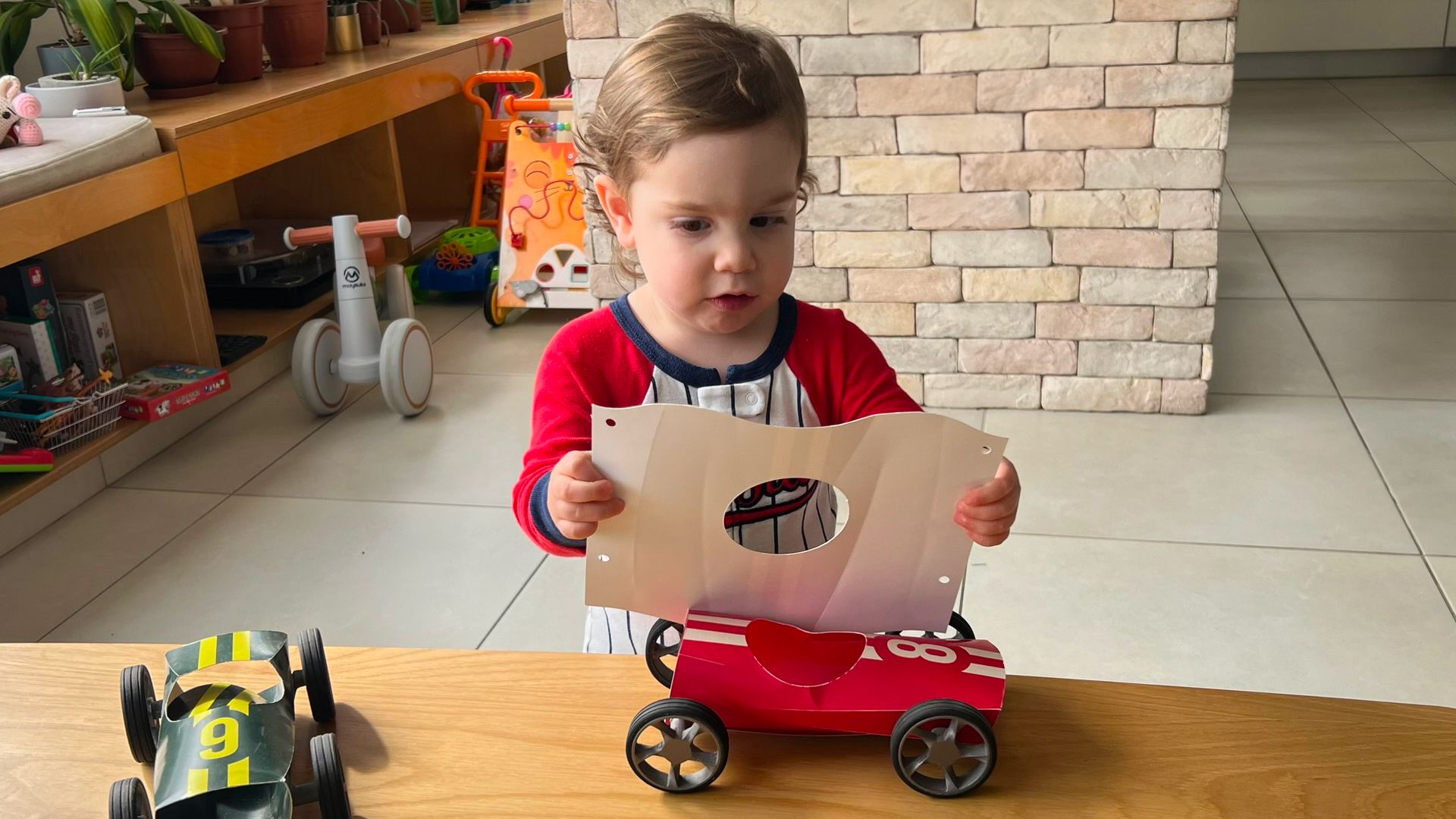 A toddler unfolding a flat Bendit cardboard sheet, an assembled red car on the table.