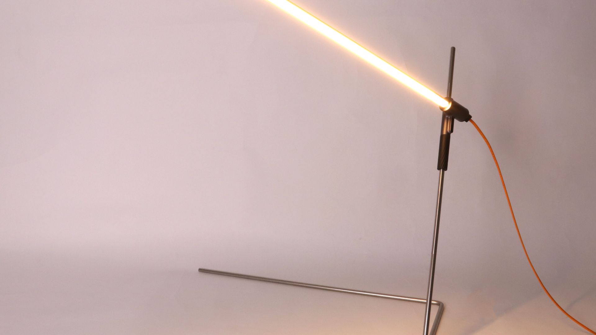 The fully assembled Rodlight lamp switched on — a slim minimal frame with a glowing LED bar and orange cable on its stand.